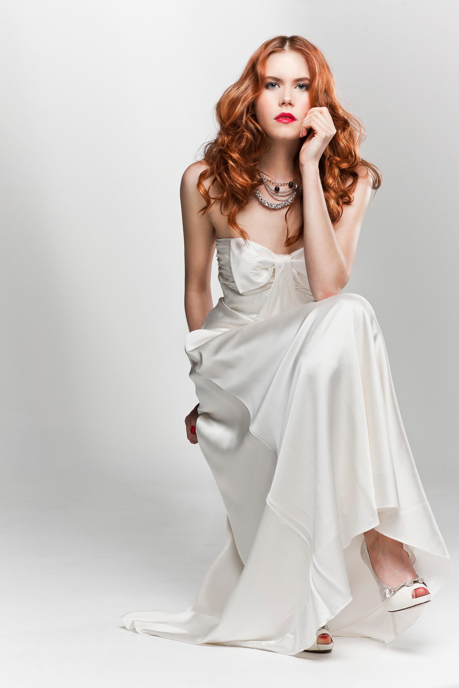 Studio fashion photography of seated woman with long red wavy hair wearing long white dress